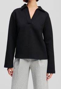 Polo de manga larga negro en tejido suave con escote en V y cuello, que presenta un sutil logo en el pecho y un corte relajado.