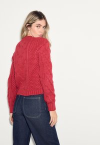 Rode kabelgebreide trui met een kort ontwerp en geribbelde manchetten. Draagt u met donkerblauwe jeans, voorzien van zakken en een ontspannen pasvorm.