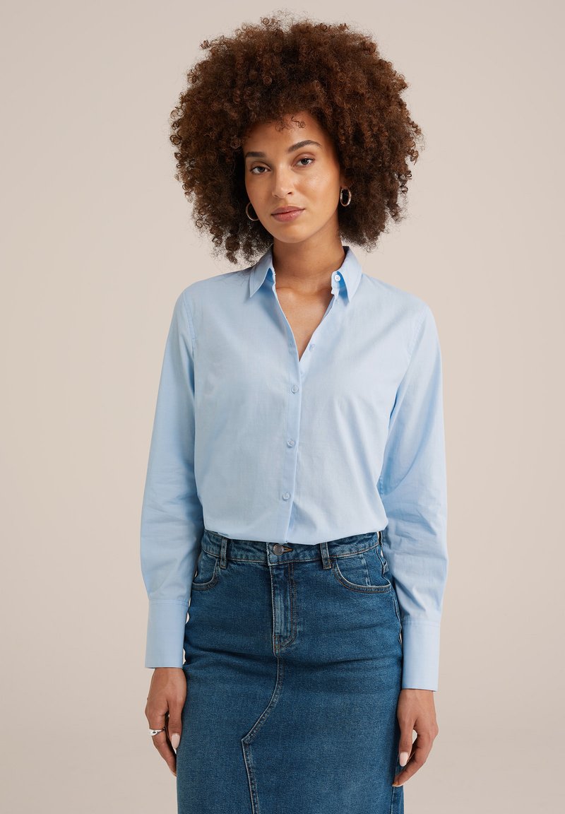 Donna con capelli ricci che indossa orecchini a cerchio argentati, camicia azzurra con bottoni e gonna di jeans scura a vita alta su sfondo semplice.
