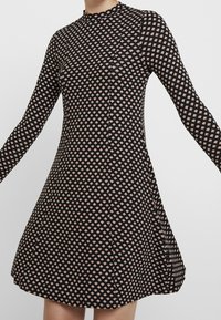 Svart och beige långärmad klänning med hög halsringning och geometriskt diamantermönster, åtsittande vid midjan och utställd vid kjolen.