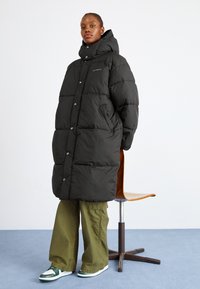 Zwarte puffer jas met een hoge kraag, drukknoopjes en zijzakken. Draagt over olijfgroene wijde broeken en witte sneakers met groene accenten.