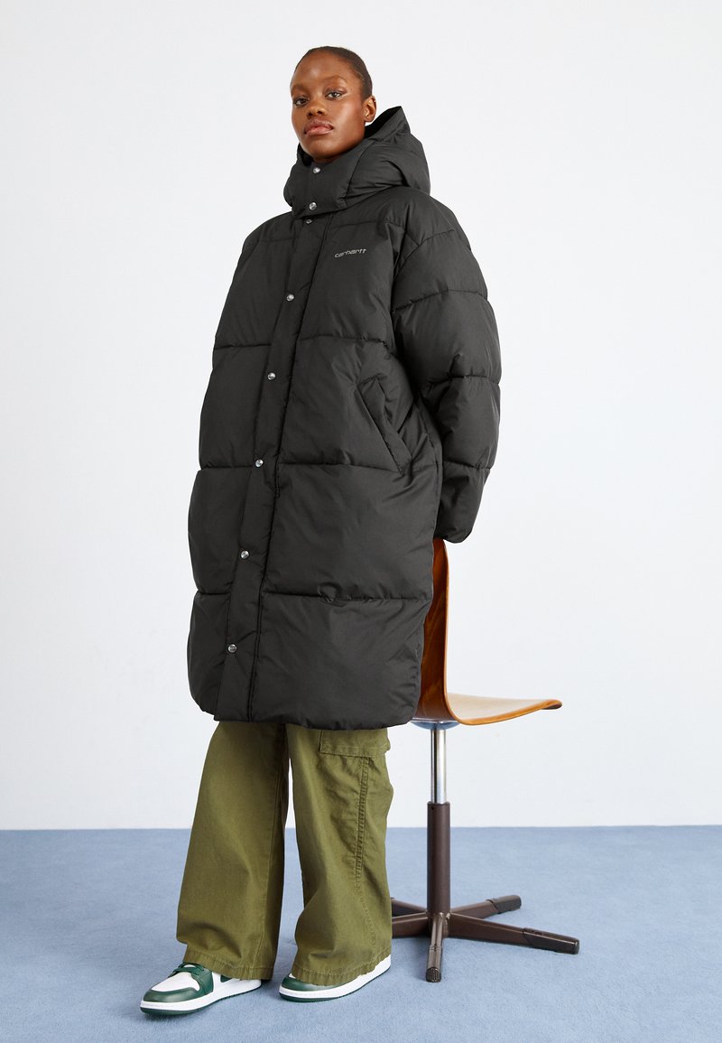 Zwarte puffer jas met een hoge kraag, drukknoopjes en zijzakken. Draagt over olijfgroene wijde broeken en witte sneakers met groene accenten.