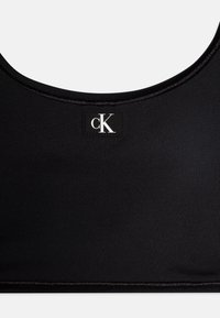 Juoda sportinė liemenėlė, pagaminta iš гладkios medžiagos, su apvaliu iškirpimu ir maža balta "cK" logotipo etikete priekyje.