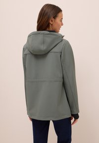 Veste à capuche verte avec une texture lisse, dotée d'une couture renforcée sur le dos, associée à un jean bleu foncé.