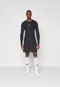 Sort langærmet sportstop med hvidt logo, sorte shorts, hvide sokker med sorte striber og hvide fodboldstøvler. Enkelt, tætsiddende design.