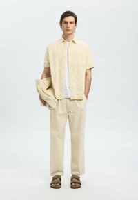 Camicia button-up beige chiaro sopra una maglietta bianca, abbinata a pantaloni beige e sandali verde scuro. Il tessuto ha una leggera texture.