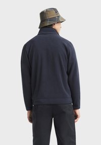 Pullover in pile di un profondo blu scuro con colletto alto; texture morbida; abbinato a un cappello a secchiello a quadri nelle tonalità marrone e blu.