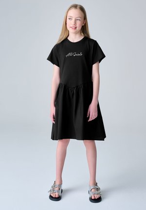 Chica con largo pelo rubio, vestida con un vestido negro de manga corta con texto "All Saints" y sandalias plateadas con hebilla, de pie sobre un fondo liso y claro.