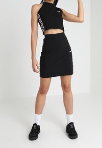 Černý zkrácený halter top a přiléhavá tužková sukně s logo akcenty v bílé a červené, doplněné černými teniskami a bílými ponožkami.