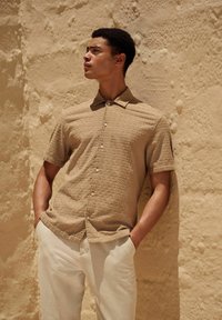 Chemise à manches courtes en tissu marron clair texturé, associée à un pantalon crème, sur un fond neutre.