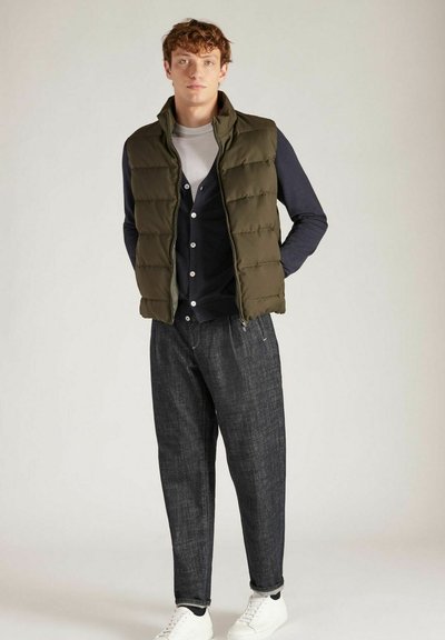 Gilet rembourré vert olive sur un pull sombre avec des boutons, assorti à un pantalon sombre et des baskets blanches. Le gilet a un col montant et un design matelassé.