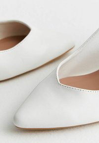 Pár bílých kožených balerín s špičatou špičkou a nízkým vykrojením v béžovém interiéru na světlém pozadí.