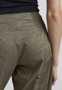 Pantalon vert olive avec un détail de taille smockée et une étiquette PULZ JEANS au dos, confectionné dans un tissu lisse et léger.