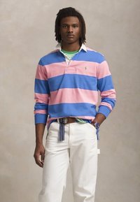 Tricou de rugby cu mânecă lungă, cu dungi orizontale roz și albastre, guler alb și un mic logo. Purtați peste un tricou cu dungi verzi și asociat cu pantaloni albi.