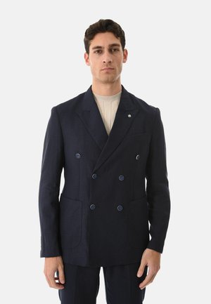 Uomo che indossa un blazer doppiopetto blu scuro sopra un maglione a coste beige chiaro, in piedi davanti a uno sfondo chiaro e semplice.