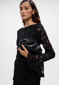 Melnas ziedu mežģīņu kleitas ar caurspīdīgām garām piedurknēm, ar paplatinātām aprocēm; tur melnu ādas somiņu ar sakumpušu detaļu.