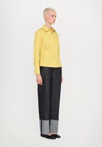 Chemise jaune à boutons avec manches longues ; associée à un pantalon large en denim foncé avec des poignets gris et des chaussures noires. Design minimaliste.
