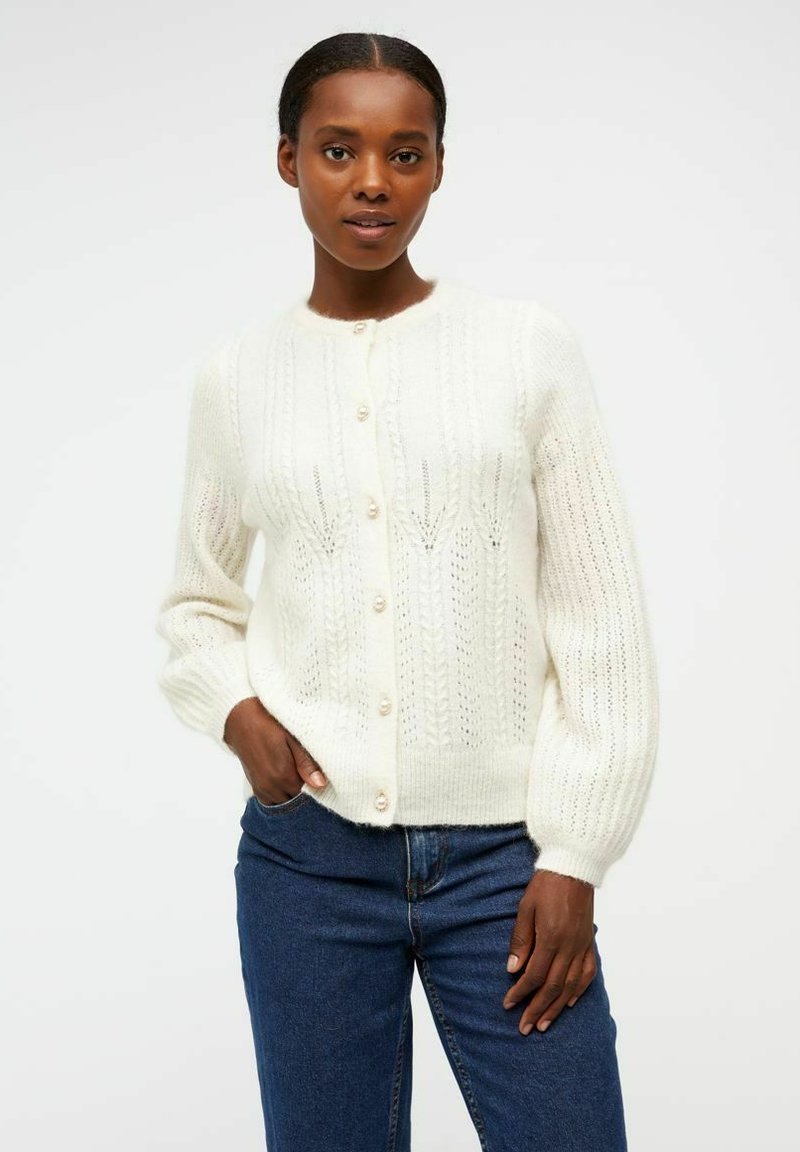 Cardigan en tricot blanc avec un col rond, fermeture à boutons et motif texturé. Les manches sont légèrement bouffantes, ajoutant de la dimension.