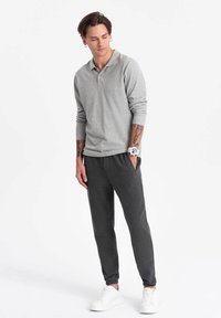 Camisa polo de manga larga gris claro, de tejido suave, combinada con pantalones jogger gris oscuro. El modelo lleva zapatillas blancas y tiene tatuajes visibles.