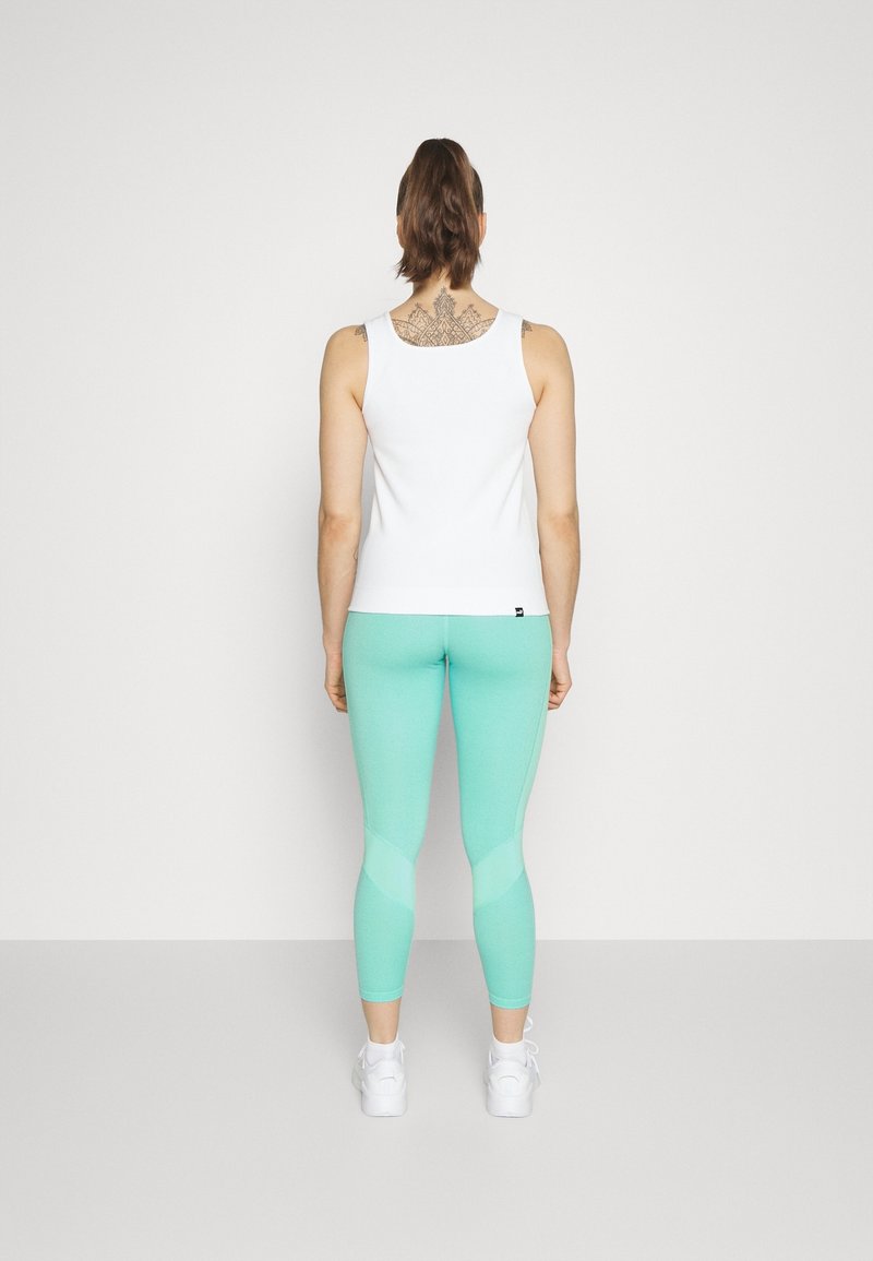 Kobieta z kucykiem, ubrana w białą bezrękawnik, miętowe legginsy, białe trampki i ozdobny tatuaż na górnej części pleców, odwrócona tyłem.