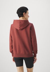 Bordó kapucnis hoodie, állítható kapucnival, bordás ujjak és szegély, puha anyagból készült. A modellen fekete rövidnadrág van, amely a laza, kényelmes stílust tükrözi.