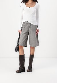 Valkoinen urheilu pitkähihainen paita, jossa on musta nauha, harmaat polvimittaiset shortsit ja ruskeat nahkaiset polvipituiset saappaat soljilla, pitäen mustaa laukkua.