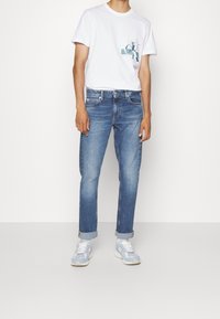 Camiseta blanca de algodón con logo gráfico, combinada con unos vaqueros de denim azul de corte recto y puños doblados, acompañada de zapatillas de deporte azul claro.