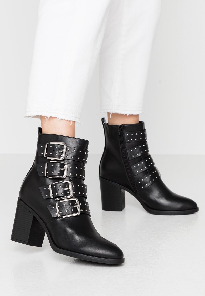 Botas de tobillo negras de cuero suave con cuatro correas que tienen grandes hebillas plateadas y acentos de tachuelas, y un tacón bloque.