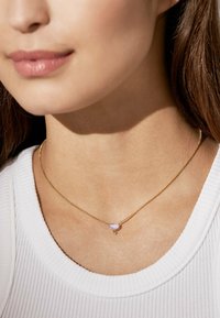Goldene Halskette mit einem kleinen, helllila Tropfenanhänger. Getragen zu einem strukturierten weißen Oberteil, das die Farbe und Form des Anhängers hervorhebt.