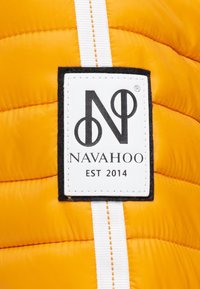 Geel pufferjack met een gestikte textuur, voorzien van een wit en zwart label met de tekst "NAVAHOO" en "EST 2014."