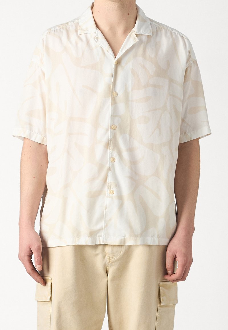 Uomo che indossa una camicia a maniche corte con bottoni, caratterizzata da un sottile motivo di foglie tropicali beige, e pantaloni cargo beige chiaro.