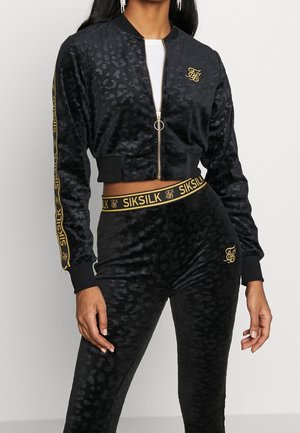 Kvinde iført sort fløjlsagtig cropped jakke og matchende højtaljede bukser med subtilt leopardprint og gyldne SikSilk-logo-detaljer.