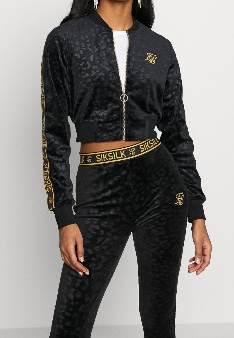 Femme portant une veste courte veloutée noire et un pantalon taille haute assorti avec un imprimé léopard subtil et des accents dorés de la marque SikSilk.