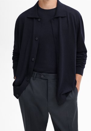 Homme portant un cardigan noir boutonné, une chemise noire à manches longues et un pantalon habillé sombre, les mains dans les poches sur fond blanc.