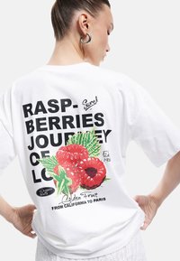 Camiseta de algodón blanca con un estampado colorido de frambuesas, texto en negrita negro y un cuello redondo, con mangas cortas y un corte holgado.