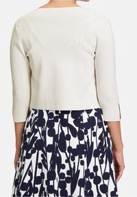 Avkortad gräddfärgad tröja med trekvartsärmar, slät textur och öppna sidor, bärs över en mönstrad kjol i marinblått och vitt.