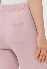 Close-up van de onderlichaam van een persoon die een lichtroze broek draagt met een elastische tailleband en een achterzak, met een hand naast de heup.