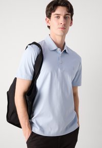 Camisa polo de manga curta azul clara com colarinho, placket de dois botões e logótipo bordado. Saco tote preto pendurado no ombro.