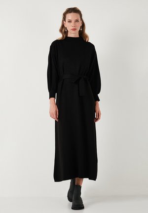 Schwarzes, langärmeliges Kleid mit hohem Kragen und einem Stoffgürtel. Hergestellt aus weichem Material, mit lockerer Passform und subtiler Textur. Schwarze Stiefeletten.