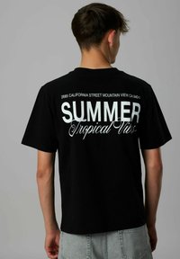 Junger Mann trägt ein schwarzes T-Shirt mit weißem Text "2685 California Street Mountain View CA 94040 SOMMER Tropische Vibes" auf dem Rücken.