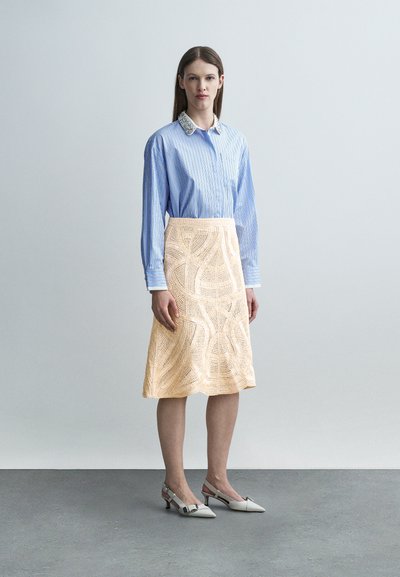 Femme portant une chemise bleue à rayures avec un col décoratif, une jupe beige à motifs longueur genou, et des chaussures blanches à talons bas avec bride arrière, debout.