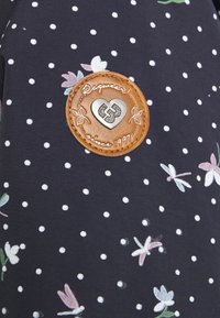 Marinfärgad tyg med vita prickar och blommönster. Har en brun läderlapp med präglad hjärtformad design och texten "Raqueta sedan 1997."