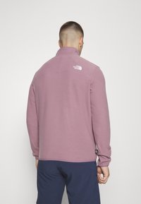 Fleece pullover i lyse lilla med høj krave, lange ærmer og et hvidt logo på ryggen. Glat tekstur uden synlige mønstre.