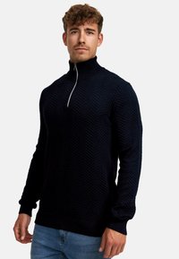 Marineblauer Strickpullover mit hohem Kragen und halbseitigem Reißverschluss, der ein strukturiertes Fischgrätenmuster und lange Ärmel aufweist.