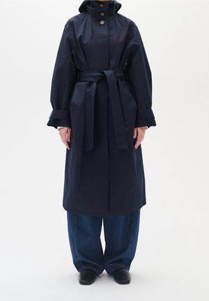 Trench-coat bleu marine à ceinture avec capuche et fermeture à boutons, porté par-dessus un jean ample foncé et des chaussures noires sur un fond blanc uni.