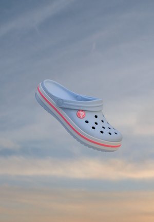Un sabot Crocs bleu clair, doté de trous d’aération et d’une bande rose, flotte devant un ciel nuageux.