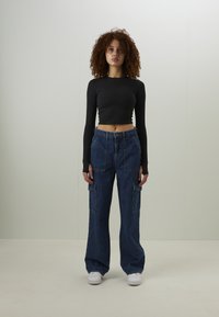 Top preto cropped de mangas longas combinado com calças cargo de denim largas com dois bolsos frontais. Modelo usa ténis brancos. Fundo neutro.