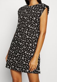 Robe noire à fleurs avec de courtes manches à capuchon. Le matériau est léger, avec un motif floral blanc dispersé. Silhouette droite, longueur genou.