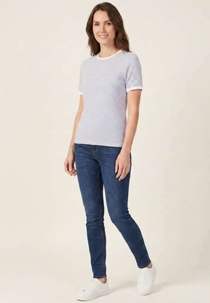 Femme debout, portant une chemise à manches courtes blanche et gris clair rayée, un jean bleu et des baskets blanches, sur un fond uni.