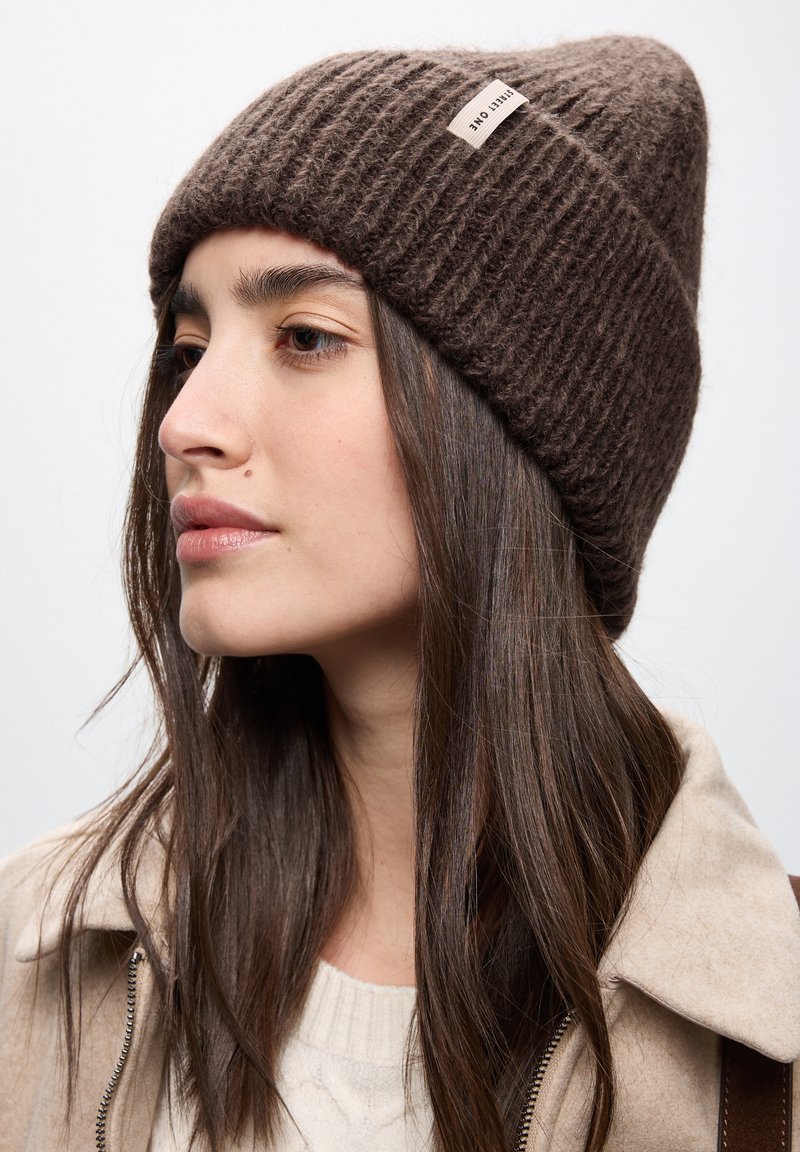 Cappello di lana marrone a maglia con texture a coste, punta appuntita e una piccola etichetta con logo sul lato. Indossato da un modello con capelli lunghi e scuri.